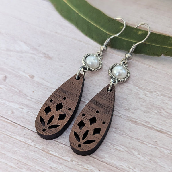 A pair of handmade teardrop wooden dangle earrings with laser-cut leaf patterns, featuring silver-toned metal hoops with white faux pearls, displayed on a light wood surface next to a green leaf.