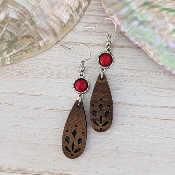 A pair of bohemian-style wooden dangle earrings with laser-cut floral patterns, red stones, and silver-tone hook wires, displayed on a light wood surface.