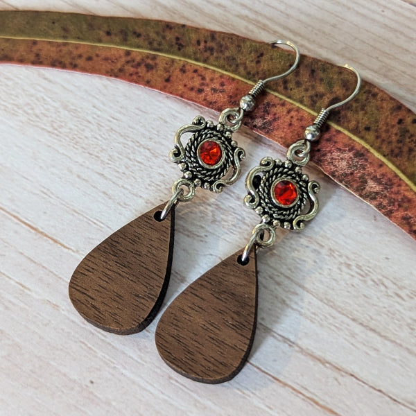 A pair of handmade dangle earrings with teardrop-shaped dark brown wooden pendants, ornate antique silver filigree tops, and round red centers, displayed on a light wood surface with a curved decorative leaf.