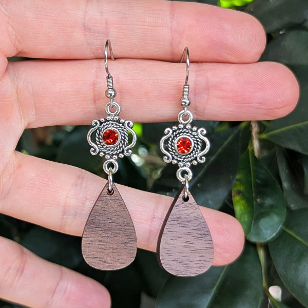 A pair of handmade dangle earrings held in a hand, featuring silver filigree tops with round red crystal and teardrop-shaped dark wood pendants, set against a background of green leaves.