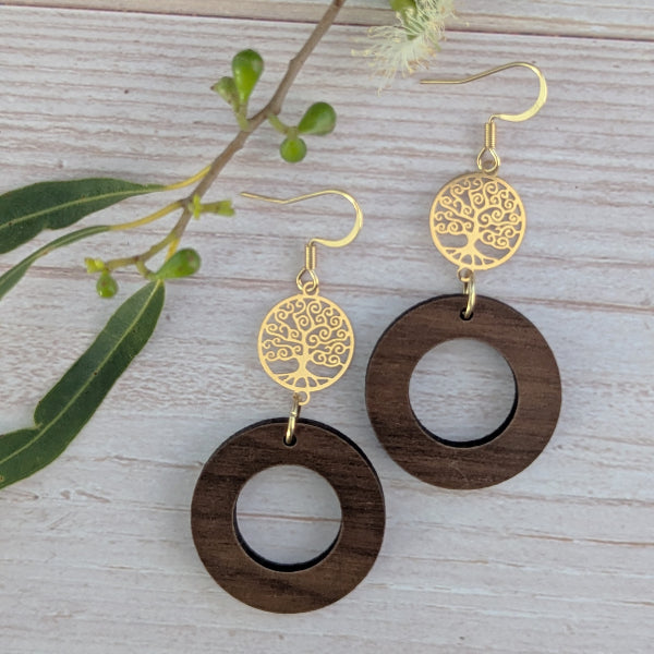 Wooden earrings with gold tree-of-life design on a wooden surface with leaves.