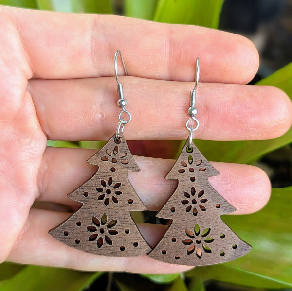 Wooden Christmas tree-shaped earrings with floral cutouts held in a hand against a green leafy background. Stainless steel hooks.