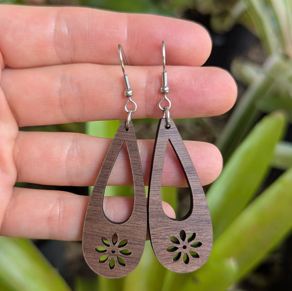 Wooden teardrop earrings with floral cutouts held between fingers against a green leafy background. Stainless steel hooks.