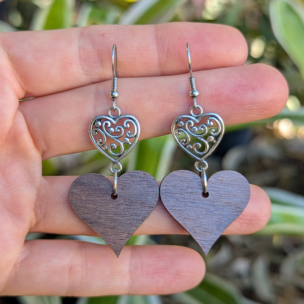 Heart-shaped wooden earrings with silver filigree details held on a hand. Stainless steel hooks.