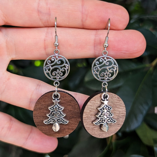 A pair of handmade drop earrings held in a hand, featuring circular wooden pendants with silver tree motifs and ornate silver filigree circles above, set against a background of dark green leaves.