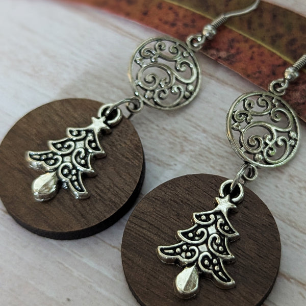A pair of handmade dangle earrings featuring ornate silver-toned filigree circles and Christmas tree charms, suspended from round dark brown wooden discs, displayed on a light wood surface.