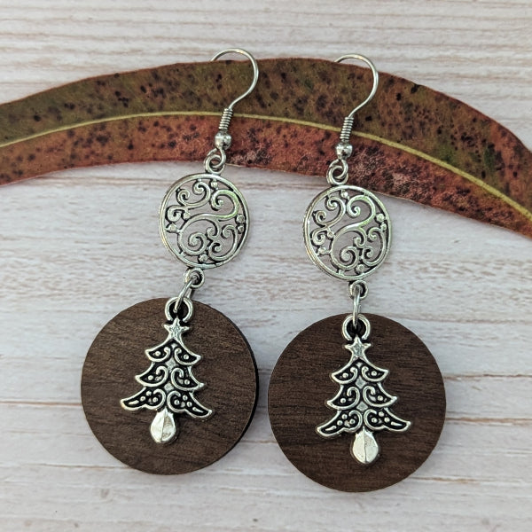 A pair of handmade dangle earrings featuring oval dark brown wooden discs, silver filigree circular charms, and silver Christmas tree pendants, displayed on a light wood background with a decorative leaf.
