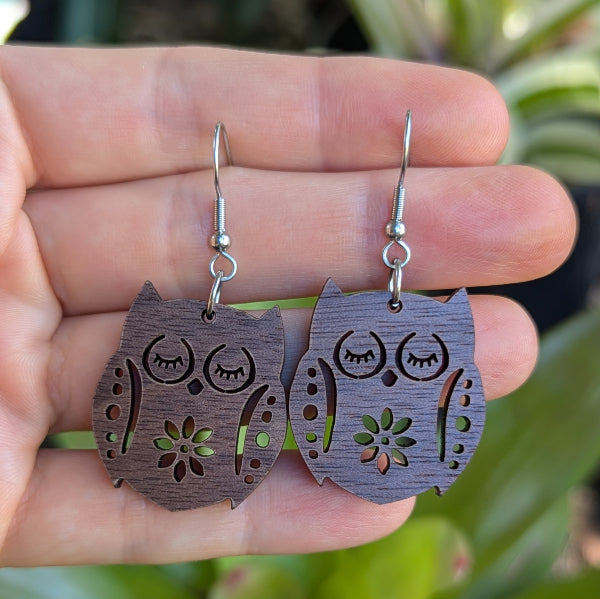 Wooden owl earrings held between fingers with a blurred natural background. Stainless steel hooks.