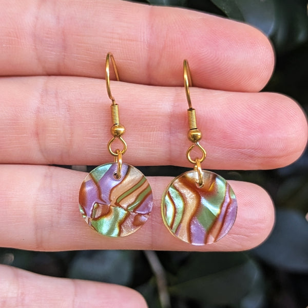 A pair of gold-tone dangle earrings with round acrylic disc pendants, held between fingers against a dark green leafy background.