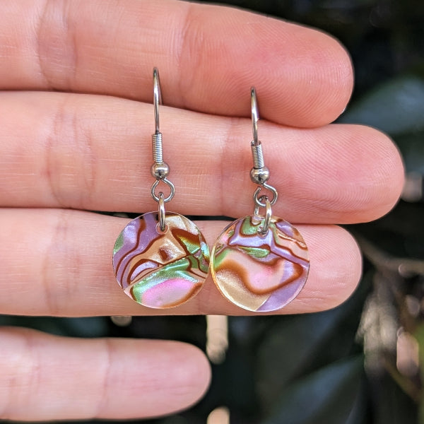 A pair of dangle earrings with iridescent acrylic disc pendants and silver fishhook wires, held in a person's hand against a blurred dark green background.