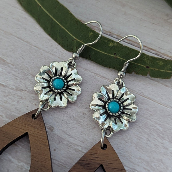A pair of silver dangle earrings with turquoise centers and black starburst detailing, displayed on a light wood surface with a green leaf and wooden cutout props.