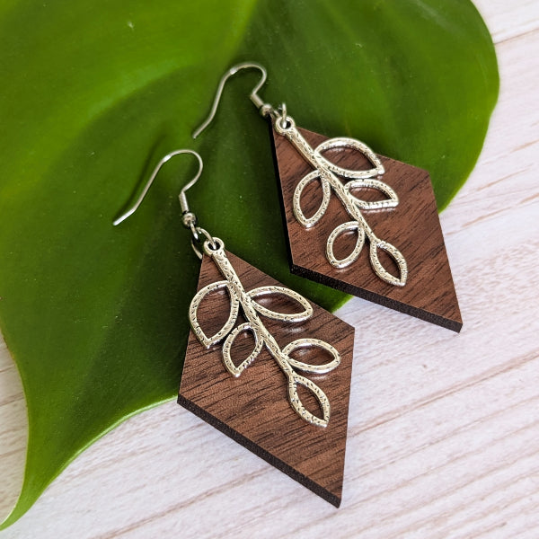 A pair of dangling earrings with dark brown wooden geometric drops and textured silver leaf motifs, displayed on a large green leaf over a light wood surface.