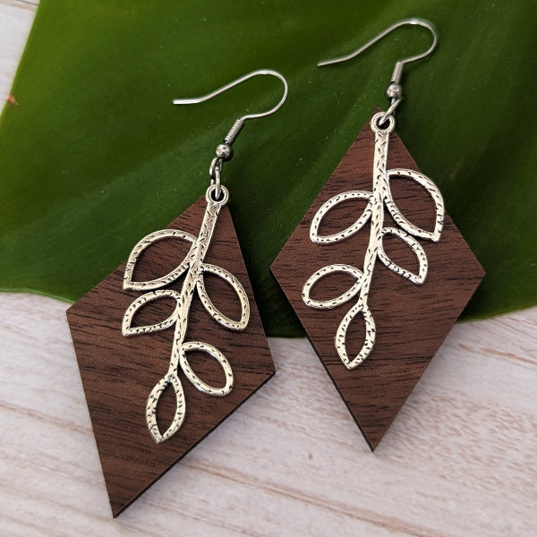 A pair of dangling earrings with dark brown wood geometric diamond-shaped pendants featuring a textured silver leaf branch design, displayed on a green leaf over a light wood surface.