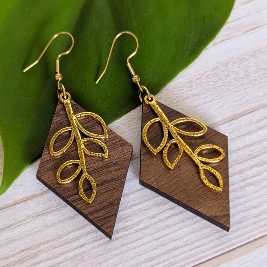 A pair of dangling earrings with dark brown wood geometric pendants and textured gold leaf filigree, displayed on a light wood surface next to a green leaf.