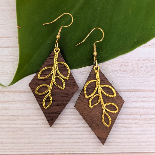 A pair of wooden drop earrings with a dark brown wood finish and a textured gold leaf design, displayed on a light wood surface next to a green leaf.