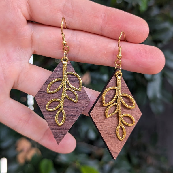 A pair of dangling earrings with dark brown wood diamond-shaped pendants and textured gold leaf motifs, held in a person's hand against a blurred green outdoor background.