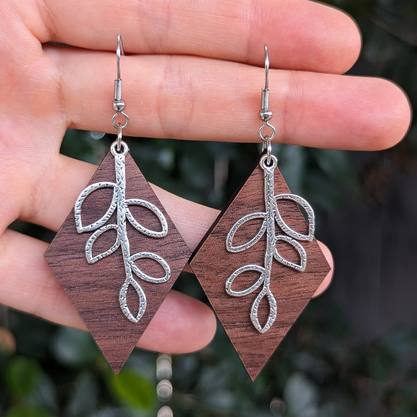 A pair of handmade dangle earrings held in a hand. The earrings feature a dark brown wood diamond-shaped drop with an oxidized silver leaf branch design, suspended from silver fishhook wires.
