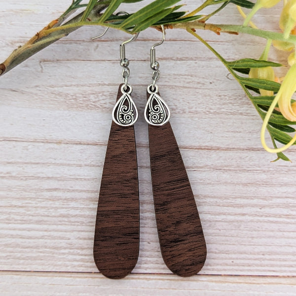 A pair of teardrop-shaped wooden dangle earrings with dark brown wood grain and silver-tone filigree details, displayed on a light wood background with green leaves and yellow flowers.