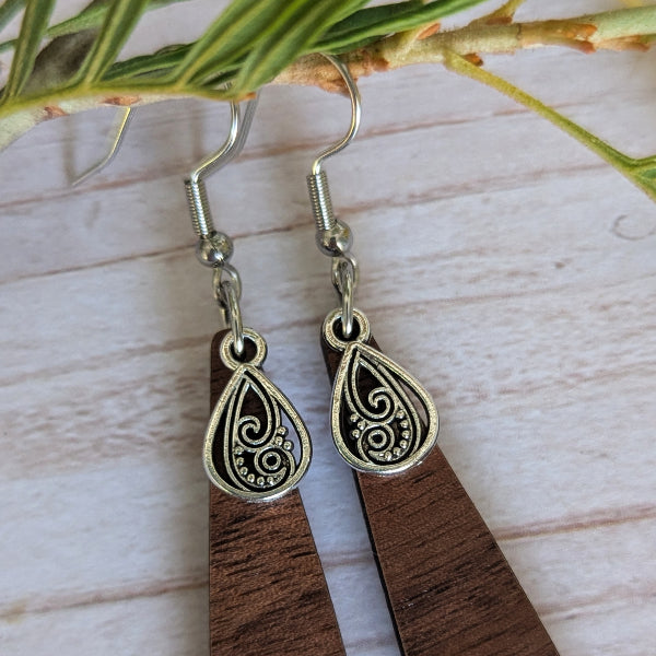A pair of handmade teardrop dangle earrings with dark brown wooden drops and ornate silver filigree tops, displayed on a light wood surface with green leaves.