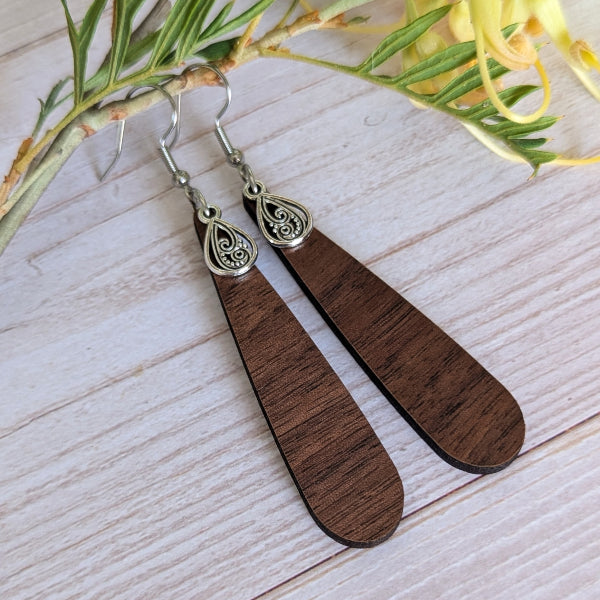 A pair of long teardrop-shaped wooden dangle earrings with silver-toned hooks and filigree details, displayed on a light wood surface with green leaves and yellow flowers as props.