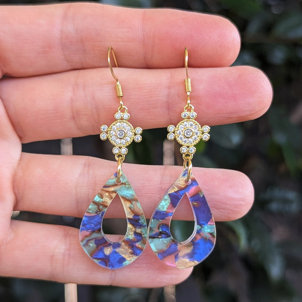 A pair of gold-tone dangle earrings with small crystal-studded floral tops and teardrop-shaped iridescent blue, purple, and gold acrylic drops, held in a person's hand against a blurred green outdoor background.