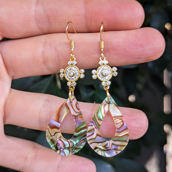 A pair of gold-tone dangle earrings with circular acrylic teardrop charms, held in a person's hand against a blurred green outdoor background.