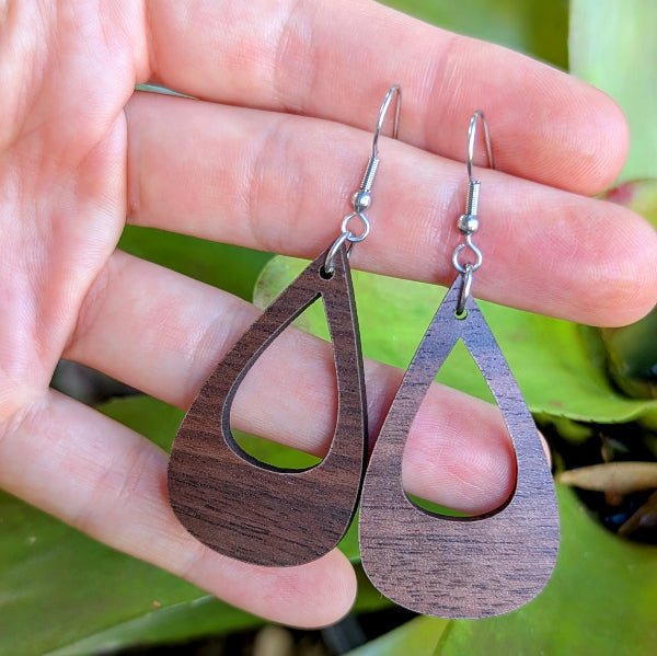 Wooden teardrop earrings held between fingers with a green leafy background. Stainless steel hooks.