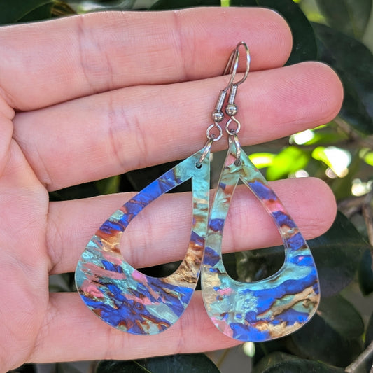 A pair of teardrop-shaped dangling earrings with a marbled blue, teal, pink, and gold pattern, held in a person's hand against a background of green leaves.