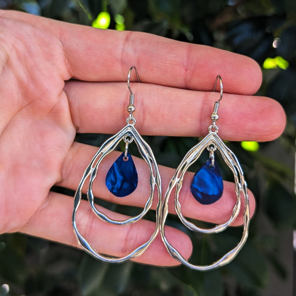 A pair of silver double-ring teardrop dangle earrings with blue acrylic inlays, held in a person's hand against a background of green foliage.