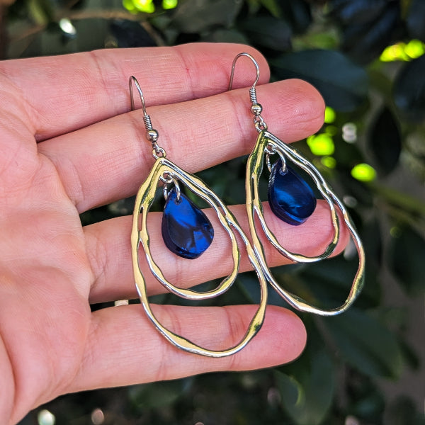 A pair of silver double-ring teardrop dangle earrings with blue acrylic inlays, held in a person's hand against a background of green foliage.