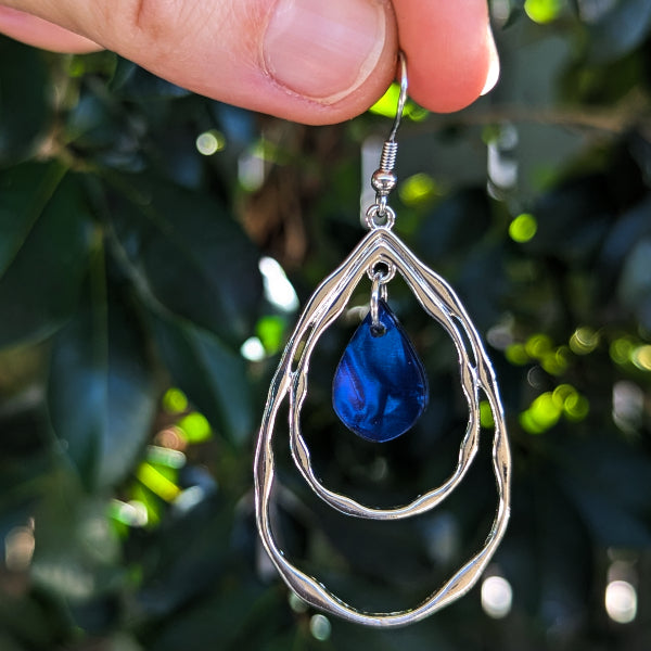 A close up of a silver double-ring teardrop dangle earring with blue acrylic inlays, held in a person's hand against a background of green foliage.