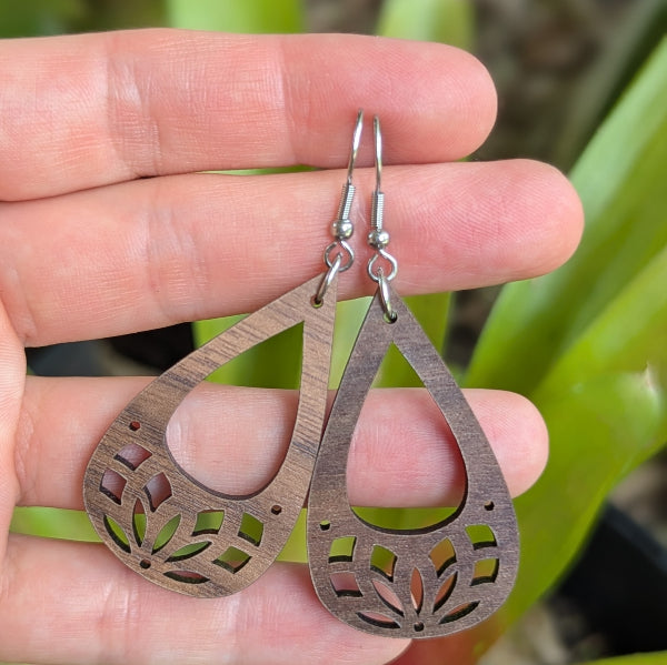 Wooden teardrop-shaped earrings held between fingers with a blurred green background. Stainless steel hooks.