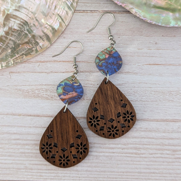 A pair of handmade teardrop earrings with laser-cut wooden drops and a colorful marbled acrylic top, displayed on a light wood surface with a seashell prop.