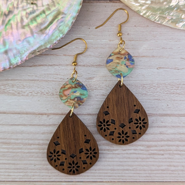 A pair of handmade teardrop dangle earrings with laser-cut wooden pendants and iridescent acrylic disc charms, displayed on a light wood background with a seashell prop.