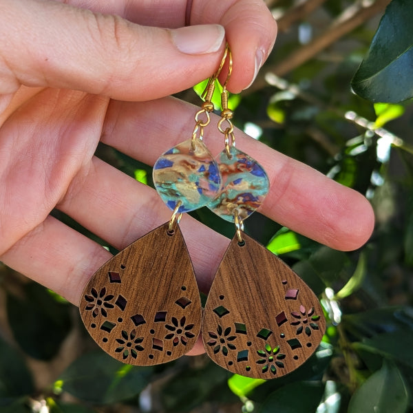 A close-up of a hand holding a pair of handmade teardrop earrings. The earrings feature a colorful marbled acrylic top half and a laser-cut wooden bottom half in a medium brown tone, with gold hooks.