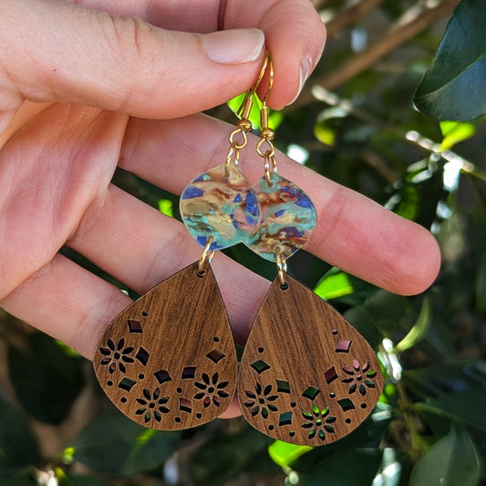 A close-up of a hand holding a pair of handmade teardrop earrings. The earrings feature a colorful marbled acrylic top half and a laser-cut wooden bottom half in a medium brown tone, with gold hooks.
