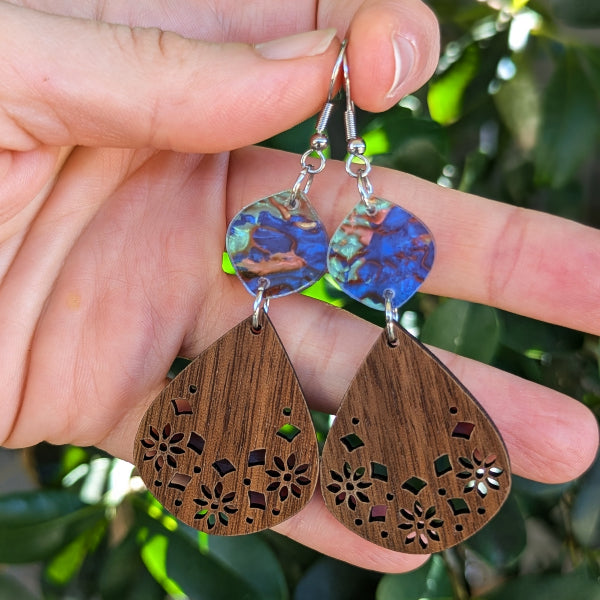 A close-up of a hand holding a pair of handmade teardrop dangle earrings. The earrings feature textured, marbled blue and green acrylic discs at the top connected to silver hook wires, and large, laser-cut wooden teardrop pendants in a medium brown wood grain with floral cutouts, set against a blurred background of green foliage.