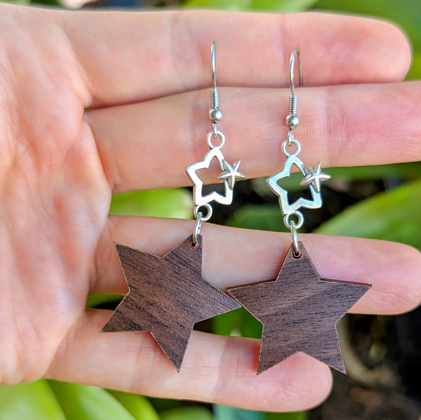 Wooden star earrings held between fingers with a blurred green background. Stainless steel hooks.