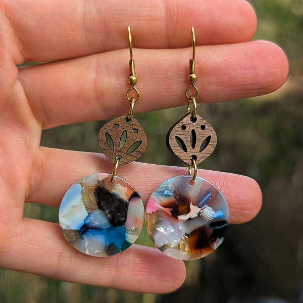 A pair of handmade dangle earrings held in a hand. The earrings feature round, marbled resin pendants with blue, white, black, and brown swirls, suspended from laser-cut wooden discs with cutout floral patterns and gold-tone hooks.