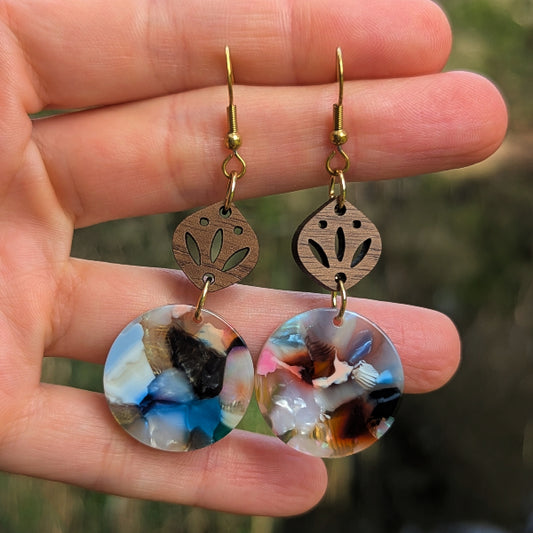 A pair of handmade dangle earrings held in a hand. The earrings feature round, marbled resin pendants with blue, white, black, and brown swirls, suspended from laser-cut wooden discs with cutout floral patterns and gold-tone hooks.
