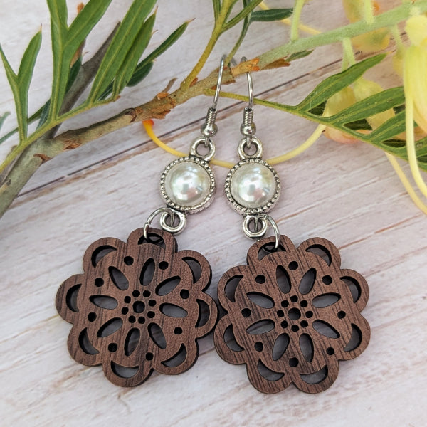 A pair of dangling earrings with oval silver-toned bezel-set white faux pearls and large dark brown wooden flower-shaped dangles, displayed on a light wood surface with green and yellow leaves in the background.