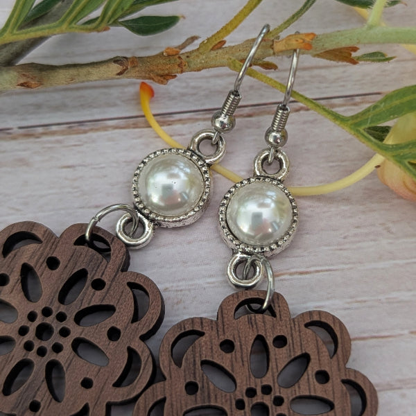 A pair of dangling earrings with round white faux pearls in silver-toned bezel settings, hanging above dark brown laser-cut wooden flower-shaped pendants on a light wood surface with green leaves in the background.