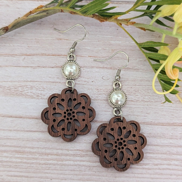 A pair of dangling earrings with silver-tone fishhook wires, round white faux pearls, and dark brown wooden flower-shaped charms with cutout floral detailing, displayed on a light wood surface with green leaves and yellow flowers as props.