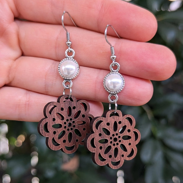 A pair of handmade dangle earrings held in a hand. The earrings feature silver-toned metal hooks and bezel-set white faux pearls, with large, laser-cut wooden dangles in a warm reddish-brown color with an intricate floral mandala pattern.