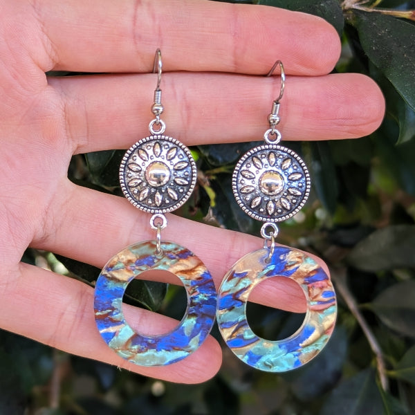 A pair of dangling earrings held in a hand against a blurred background of green leaves. The earrings feature silver-toned filigree sunburst discs and irregular circular drops with a marbled blue, gold, and turquoise pattern.