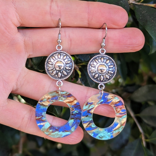 A pair of dangling earrings held in a hand against a blurred background of green leaves. The earrings feature silver-toned filigree sunburst discs and irregular circular drops with a marbled blue, gold, and turquoise pattern.