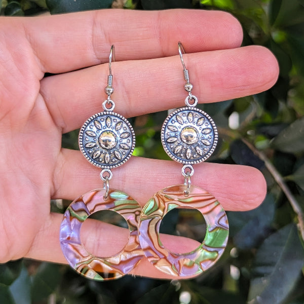 A pair of handmade dangle earrings held in a hand. The earrings feature textured silver-tone circular connectors with sunburst details and large, iridescent green, purple, and gold-toned circular drop charms.