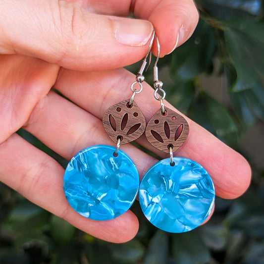 A pair of handmade dangle earrings held in a hand. The earrings feature textured bright blue circular drop charms and oval brown wooden top pieces with a cutout floral pattern, suspended from stainless steel hooks.