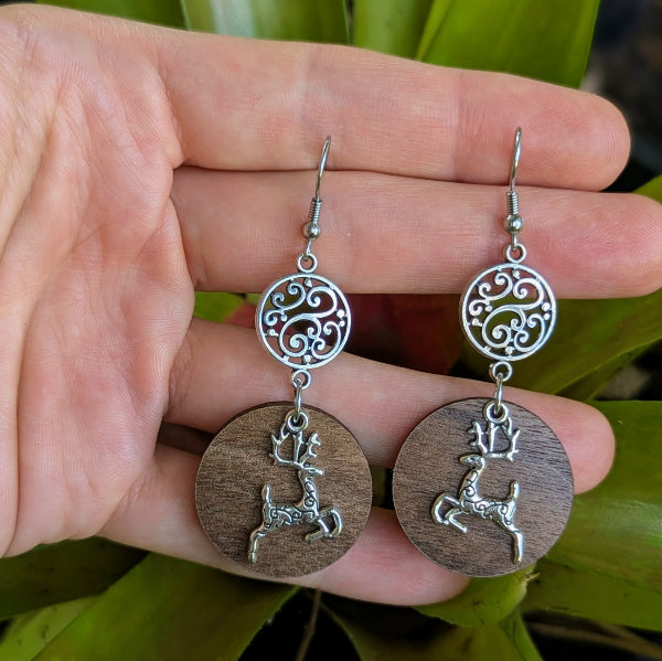 Wooden earrings with silver reindeer charms held in a hand against a green leafy background. Stainless steel hooks.