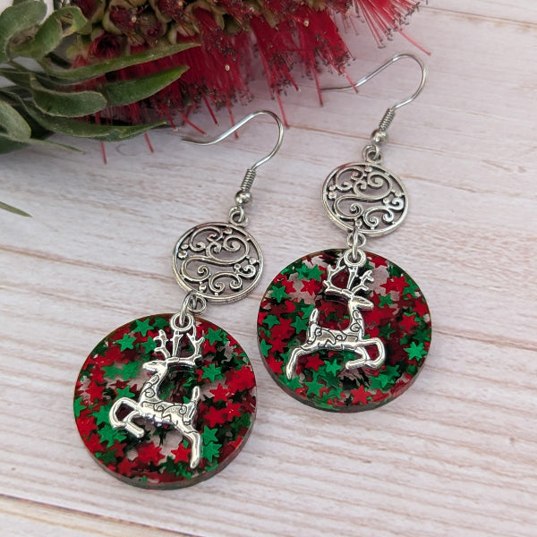 A pair of silver-toned dangle earrings with circular resin pendants featuring a red and green holiday pattern and a silver reindeer charm, displayed on a light wood surface with red flowers in the background.